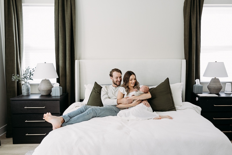 mom and dad sitting on bed holding new baby in modern room with green curtains